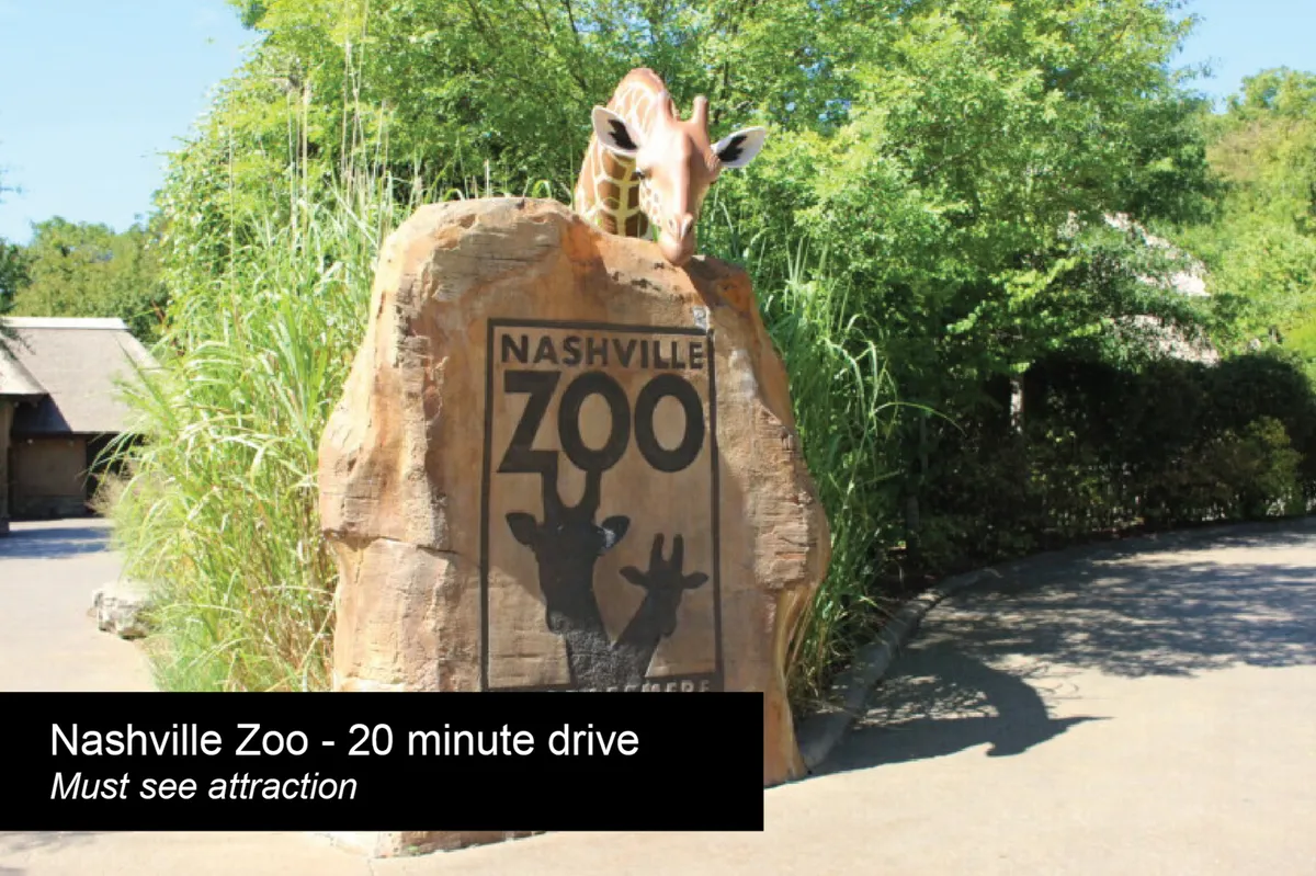 The image shows the entrance to the Nashville Zoo, featuring a large rock sign with the zoo's logo and a giraffe statue surrounded by lush greenery. Families are likely to gather here for group photos before exploring the animal exhibits.