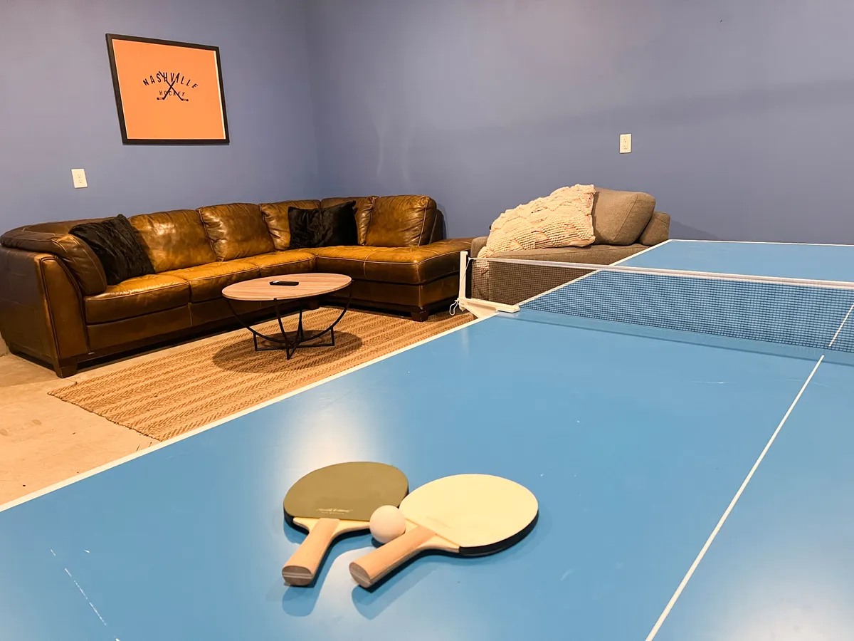 This image features a garage game room equipped with a ping pong table, leather sofa, coffee table, and attractive wall art. The scene is casual and welcoming, hinting at lively games and relaxed social time.