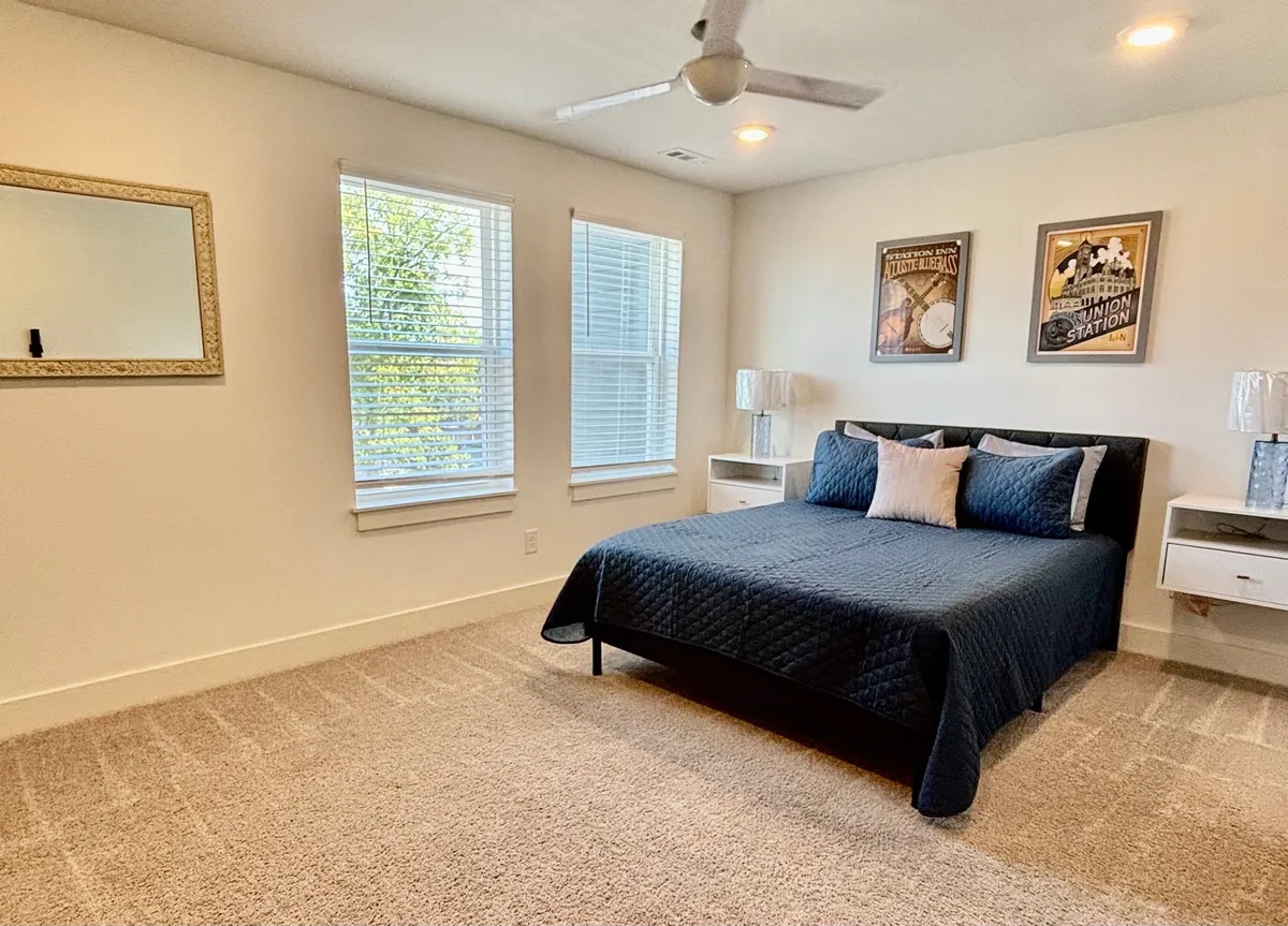 A serene bedroom with a comfortable bed, wall art, windows for natural light, a mirrored frame, and nightstands. The atmosphere is peaceful, inviting, and designed for restful moments.