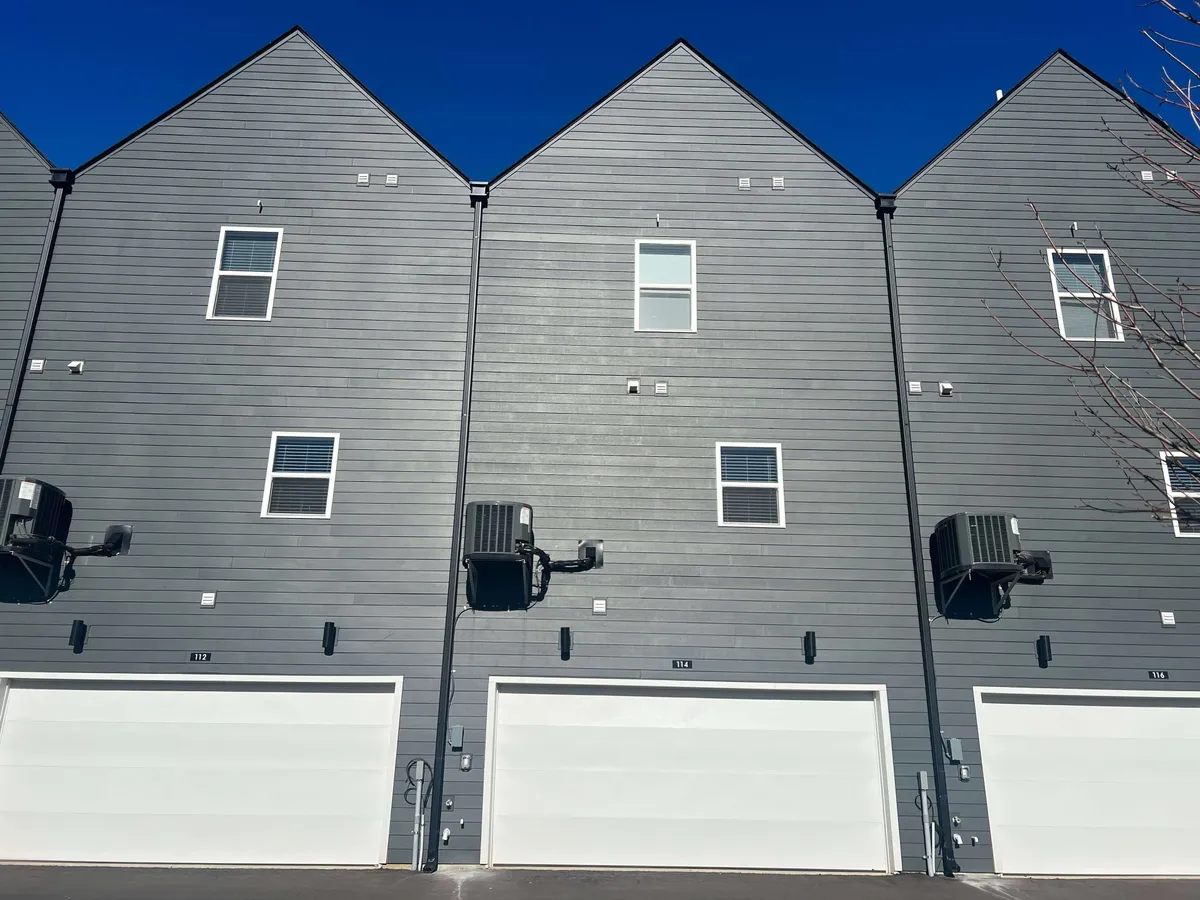 A clean, modern townhouse exterior with gray siding and white garage doors, set in a friendly, well-kept neighborhood. The image suggests multiple similar units nearby, hinting at the option to book additional homes for large groups or celebrations.